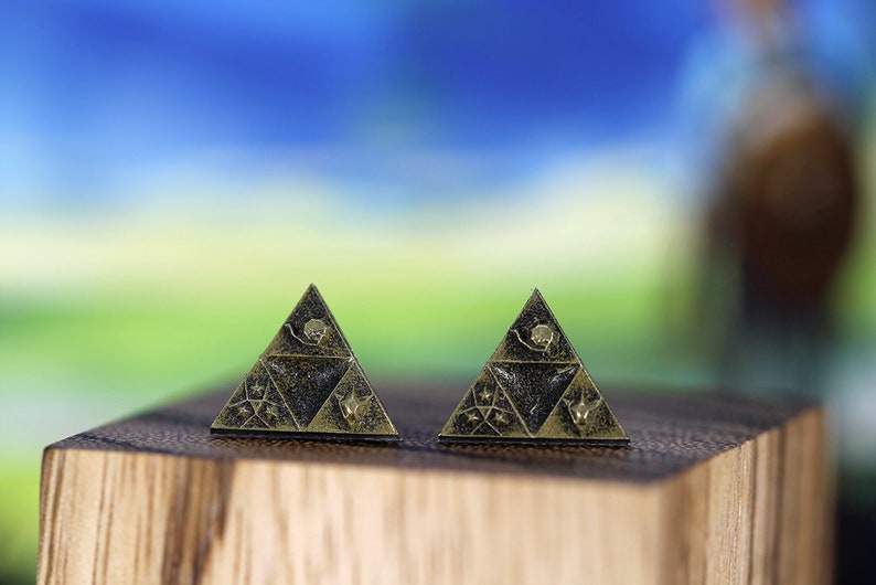 May include: Two gold-colored triangular earrings with a black background and intricate designs. The earrings are on a wooden surface.