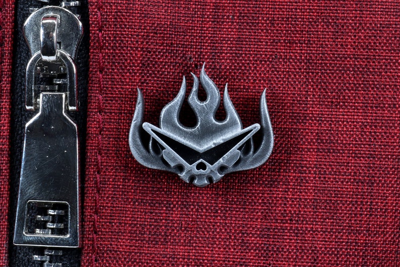 May include: A silver metal pin with a skull wearing sunglasses and a flame design. The pin is on a red fabric background.