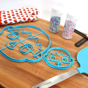 May include: Two blue plastic cookie cutters shaped like faces, a rolling pin, a spatula, two jars of sprinkles, and a black measuring tool on a wooden cutting board.