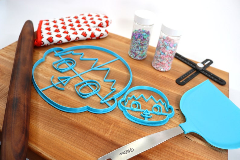 May include: Two blue plastic cookie cutters shaped like faces, a rolling pin, a spatula, two jars of sprinkles, and a black measuring tool on a wooden cutting board.