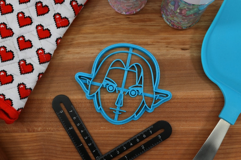 May include: Blue plastic cookie cutter shaped like Link from the Legend of Zelda video game series. The cutter is on a wooden surface.