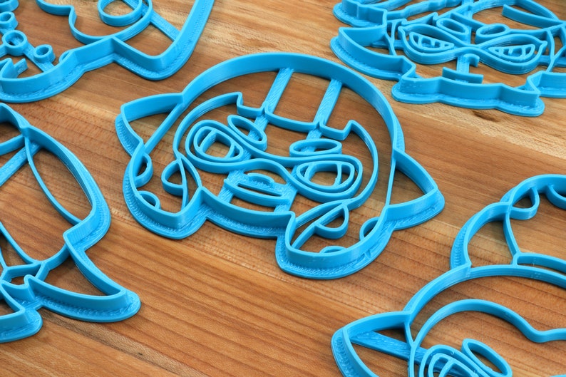 May include: A set of six blue plastic cookie cutters shaped like cartoon characters. The cutters are on a wooden surface.