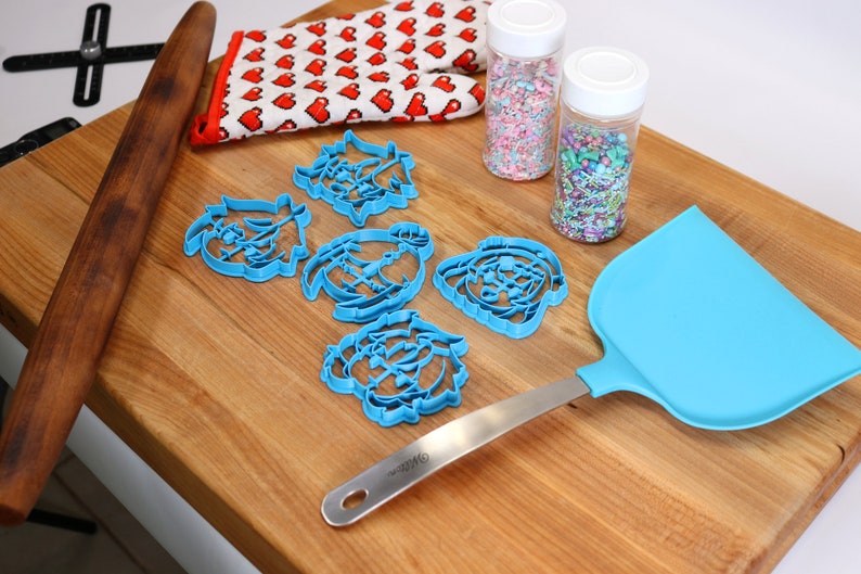 Puede incluir: Un rodillo de madera, una esp&aacute;tula azul, cinco cortadores de galletas azules, un guante de horno rojo y blanco y dos frascos de vidrio llenos de chispas de colores sobre una tabla de cortar de madera.