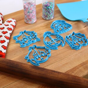 Puede incluir: Un rodillo de madera, un guante de horno rojo con corazones blancos, dos frascos de chispas, una esp&aacute;tula azul y cinco cortadores de galletas azules sobre una tabla de cortar de madera.