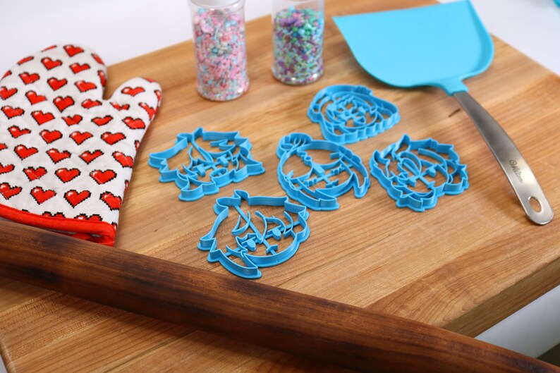 Puede incluir: Un rodillo de madera, un guante de horno rojo con corazones blancos, dos frascos de chispas, una esp&aacute;tula azul y cinco cortadores de galletas azules sobre una tabla de cortar de madera.