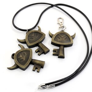 May include: Three gold-toned key-shaped pendants with a shield and horns design. The pendant on the left is attached to a black cord necklace. The pendant on the right is attached to a silver key chain.