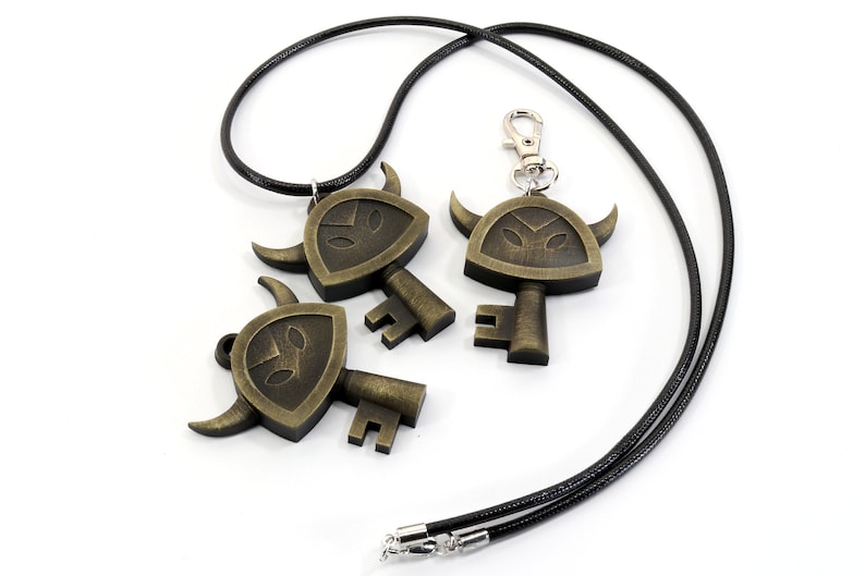 May include: Three gold-toned key-shaped pendants with a shield and horns design. The pendant on the left is attached to a black cord necklace. The pendant on the right is attached to a silver key chain.