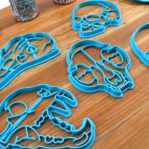 May include: Four blue plastic skull-shaped cookie cutters on a wooden surface.