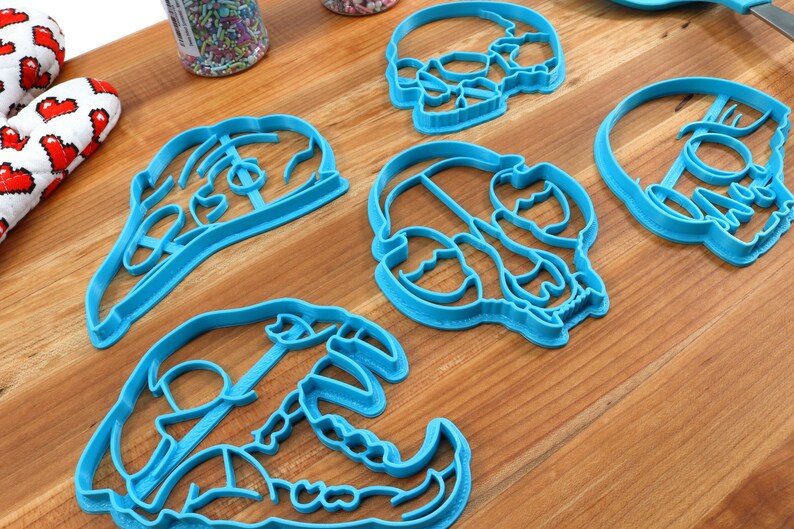 May include: Four blue plastic skull-shaped cookie cutters on a wooden surface.