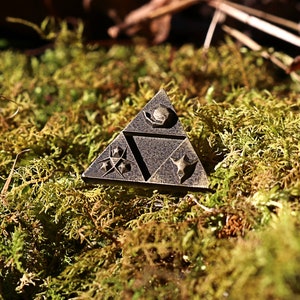 May include: A triangular metal pin with a dark brown finish. The pin features a stylized design with three symbols, each with a different shape and pattern. The pin is sitting on a bed of green moss.