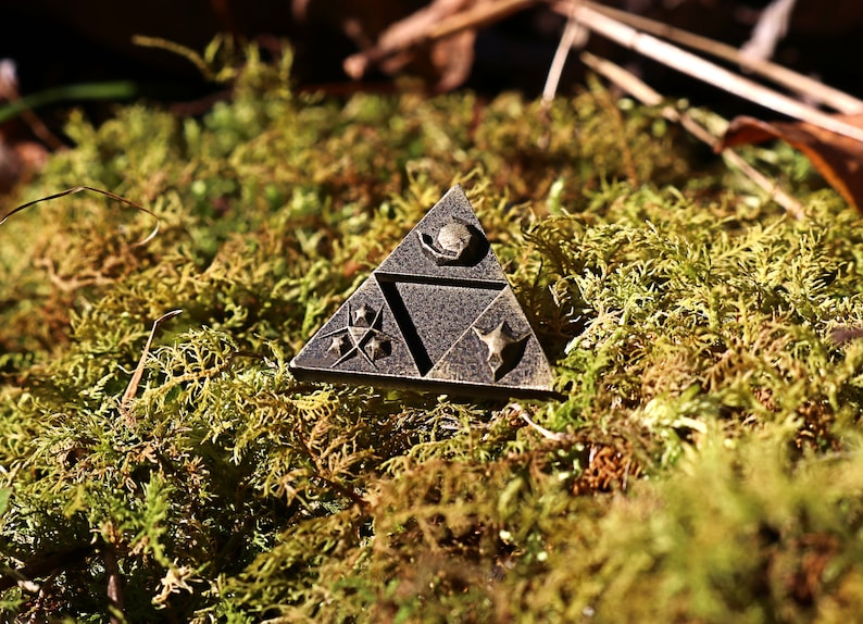 May include: A triangular metal pin with a dark brown finish. The pin features a stylized design with three symbols, each with a different shape and pattern. The pin is sitting on a bed of green moss.