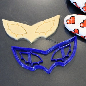 May include: A blue plastic cookie cutter shaped like a mask with two eye holes. The cookie cutter is on a dark gray surface next to a cookie that has been cut out using the cookie cutter.