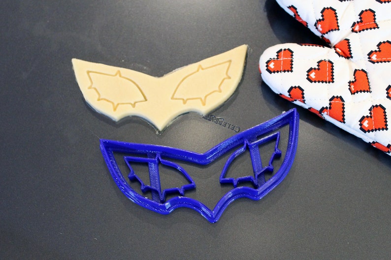 May include: A blue plastic cookie cutter shaped like a mask with two eye holes. The cookie cutter is on a dark gray surface next to a cookie that has been cut out using the cookie cutter.