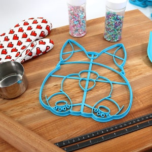 May include: A blue plastic cookie cutter shaped like a dog's head, a wooden rolling pin, a measuring cup, a red and white oven mitt, two jars of sprinkles, a black ruler, and a blue spatula on a wooden cutting board.