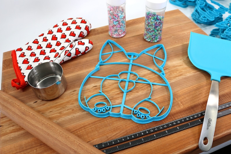 May include: A blue plastic cookie cutter shaped like a dog's head, a wooden rolling pin, a measuring cup, a red and white oven mitt, two jars of sprinkles, a black ruler, and a blue spatula on a wooden cutting board.