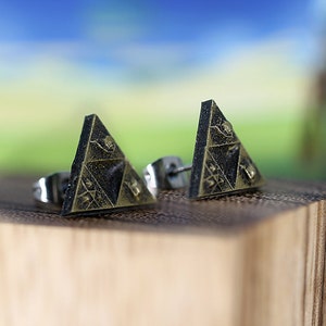 May include: A pair of gold-toned triangle earrings with a black and gold design. The earrings are on a wooden surface.