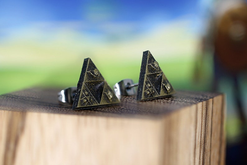 May include: A pair of gold-toned triangle earrings with a black and gold design. The earrings are on a wooden surface.