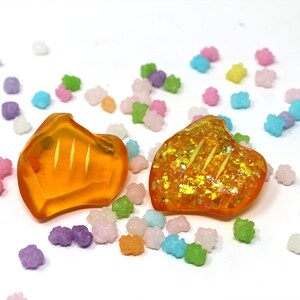 May include: Two orange resin heart-shaped earrings with gold glitter accents. The earrings are on a white background with colorful sprinkles.