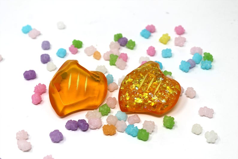 May include: Two orange resin heart-shaped earrings with gold glitter accents. The earrings are on a white background with colorful sprinkles.