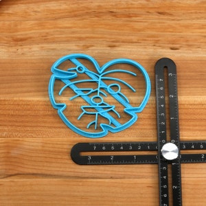 May include: A blue plastic cookie cutter shaped like a monstera leaf. The cutter is on a wooden surface next to a black measuring tool.