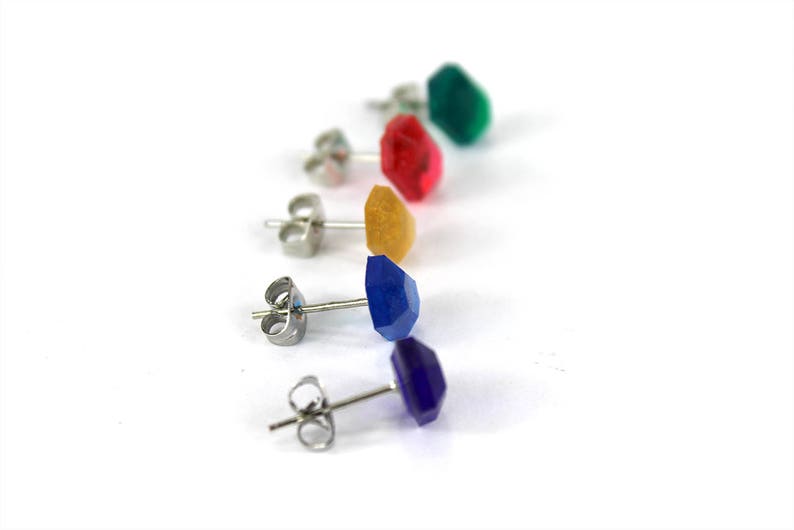 May include: Five pairs of stud earrings with different colored gemstones. The earrings are silver and have a post back. The gemstones are green, red, yellow, blue, and purple.