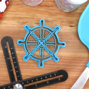 May include: A light blue plastic ship wheel-shaped cookie cutter. A black angle measuring tool is in the foreground. A blue spatula and a white cloth with red pixelated hearts are also visible. The objects are on a wooden surface.