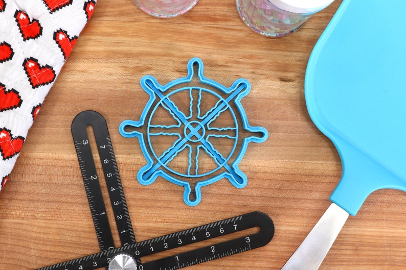 May include: A light blue plastic ship wheel-shaped cookie cutter. A black angle measuring tool is in the foreground. A blue spatula and a white cloth with red pixelated hearts are also visible. The objects are on a wooden surface.