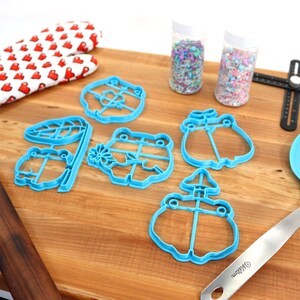May include: A set of blue frog-shaped cookie cutters, a wooden rolling pin, a pair of red oven mitts, two jars of colorful sprinkles, a blue spatula, and a black ruler on a wooden surface.