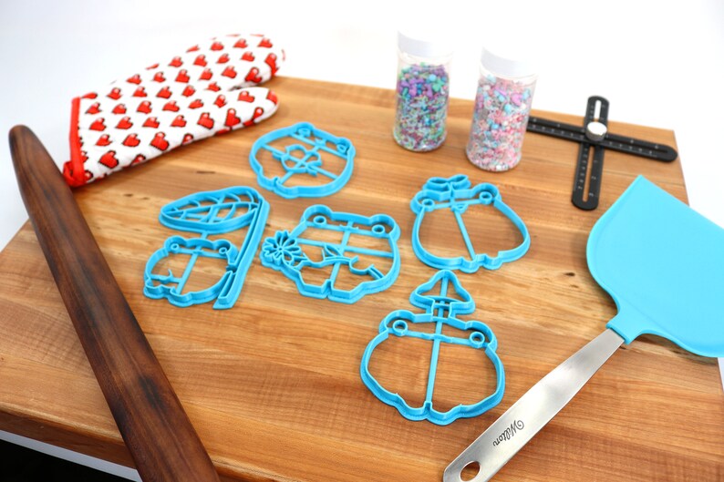 May include: A set of blue frog-shaped cookie cutters, a wooden rolling pin, a pair of red oven mitts, two jars of colorful sprinkles, a blue spatula, and a black ruler on a wooden surface.