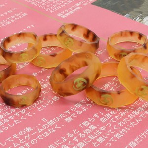 May include: A group of eight amber-colored rings with a gold design on a pink surface. The rings are made of a translucent material and have a slightly textured surface.