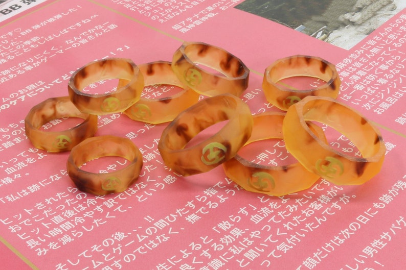 May include: A group of eight amber-colored rings with a gold design on a pink surface. The rings are made of a translucent material and have a slightly textured surface.