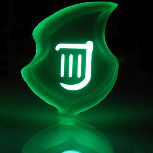May include: A luminous green shield-shaped object with a white symbol at its centre. The symbol features three vertical lines and a curved line. The object glows against a dark backdrop, producing a vibrant and striking visual.