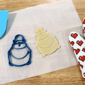 May include: A blue cookie cutter and a light brown cookie in the shape of a cartoon character on a white parchment paper surface. A red and white oven mitt is in the background.