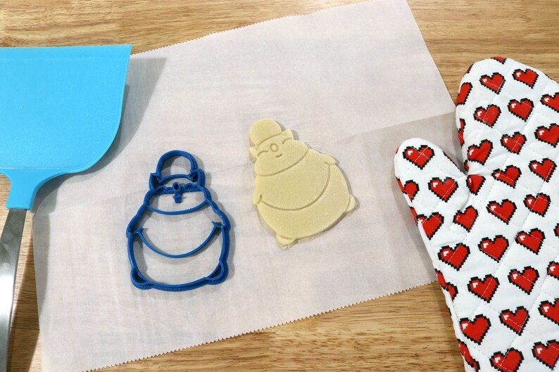 May include: A blue cookie cutter and a light brown cookie in the shape of a cartoon character on a white parchment paper surface. A red and white oven mitt is in the background.