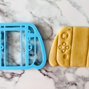 May include: A blue plastic cookie cutter shaped like a video game controller, next to a light brown cookie cut with the same shape.