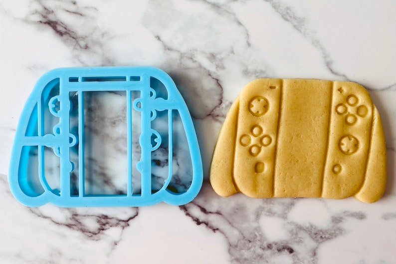 May include: A blue plastic cookie cutter shaped like a video game controller, next to a light brown cookie cut with the same shape.
