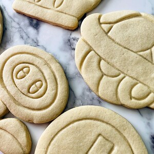 May include: Four plain sugar cookies, each with a different design. The cookies are on a white marble surface. The designs include a crown, a swirl, a circle with three dots, and a circle with a line.