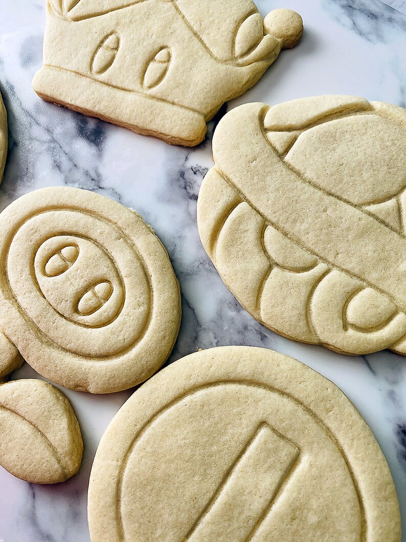 May include: Four plain sugar cookies, each with a different design. The cookies are on a white marble surface. The designs include a crown, a swirl, a circle with three dots, and a circle with a line.