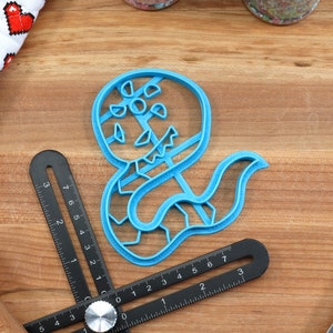 May include: A blue plastic cookie cutter shaped like a snake with a scary face. The cutter is on a wooden surface.