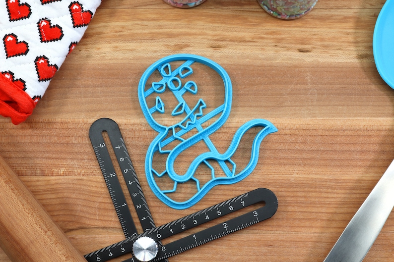 May include: A blue plastic cookie cutter shaped like a snake with a scary face. The cutter is on a wooden surface.