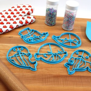 Peut inclure: Un ensemble de cinq emporte-pi&egrave;ces en plastique bleu en forme de cr&eacute;atures marines, dont une baleine, une m&eacute;duse et une narval. Les emporte-pi&egrave;ces sont sur une planche &agrave; d&eacute;couper en bois avec un rouleau &agrave; p&acirc;tisserie, une spatule et deux pots de vermicelles.