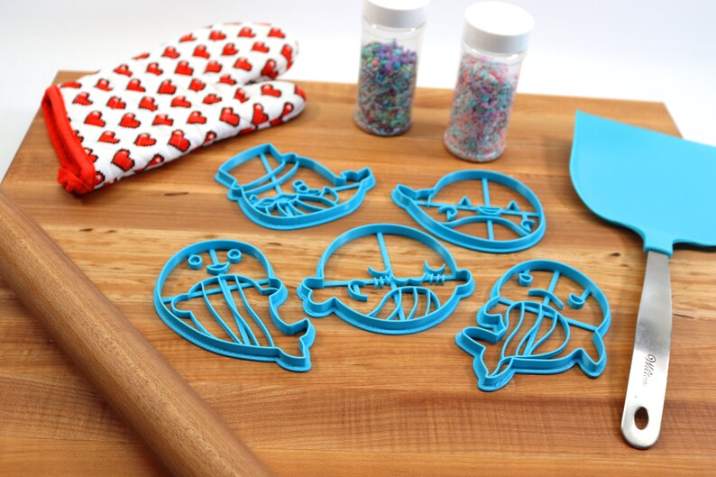 Peut inclure: Un ensemble de cinq emporte-pi&egrave;ces en plastique bleu en forme de cr&eacute;atures marines, dont une baleine, une m&eacute;duse et une narval. Les emporte-pi&egrave;ces sont sur une planche &agrave; d&eacute;couper en bois avec un rouleau &agrave; p&acirc;tisserie, une spatule et deux pots de vermicelles.