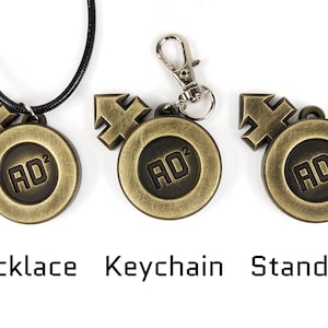May include: Three versions of a gold-colored metal pendant with a black symbol and the text "AD2". The pendant is shown as a necklace, a keychain, and a standard pendant.
