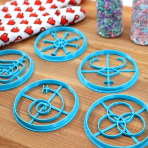 May include: Five blue plastic cookie cutters with different geometric designs on a wooden surface. The cutters are circular and feature intricate patterns.