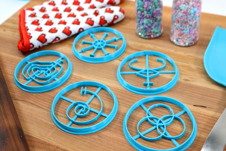 May include: Five blue plastic cookie cutters with different geometric designs on a wooden surface. The cutters are circular and feature intricate patterns.