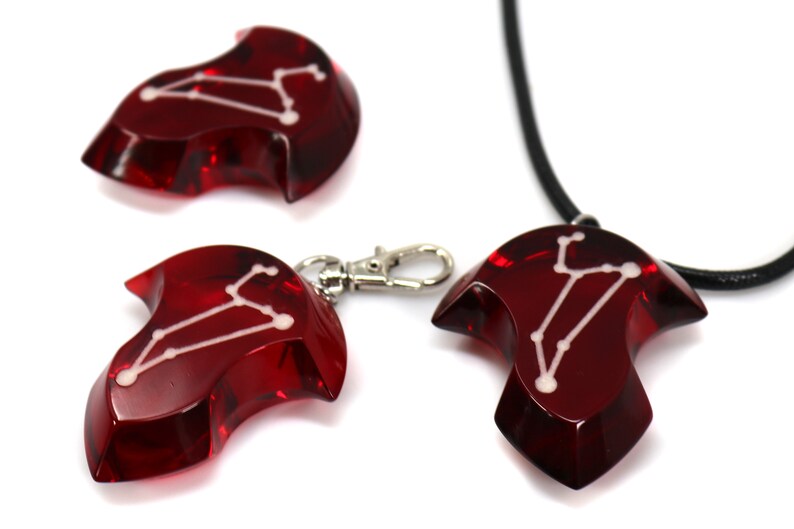 May include: Three red glass pendants with a white constellation design. The pendants are shaped like a teardrop with a pointed tip. One pendant is on a black cord necklace, one is on a silver chain, and one is loose.