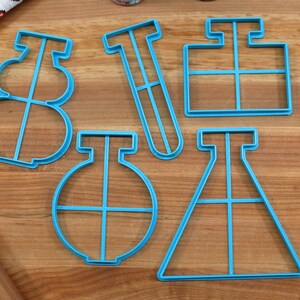 May include: Five blue plastic cookie cutters shaped like science lab equipment: a beaker, a test tube, a flask, a graduated cylinder, and a round flask. The cutters are on a wooden surface.