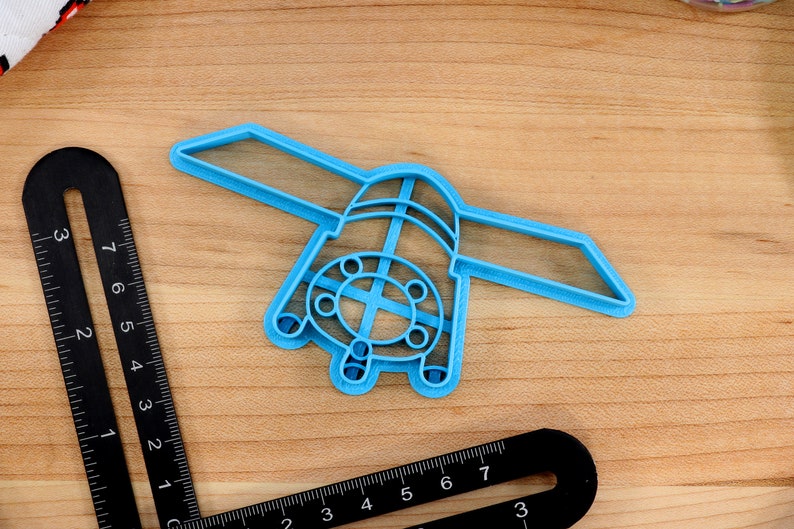 May include: A light blue plastic cookie cutter shaped like a spaceship with a round window and a cross-shaped design in the center.