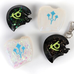 May include: Four resin keychains with different designs. Two keychains feature a green eye symbol on a black background, one with a black and gold glitter finish and the other with a green and black glitter finish. The other two keychains feature a blue trident symbol on a white background, one with a white and iridescent glitter finish and the other with a clear finish.