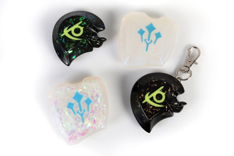 May include: Four resin keychains with different designs. Two keychains feature a green eye symbol on a black background, one with a black and gold glitter finish and the other with a green and black glitter finish. The other two keychains feature a blue trident symbol on a white background, one with a white and iridescent glitter finish and the other with a clear finish.